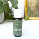 Bottle of Organic Rosemary essential oil from Oil Joy Apothecary on a white surface with green leaves in the background.