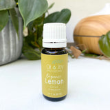 Bottle of Organic Lemon essential oil from Oil Joy Apothecary on a white surface with greenery in the background.