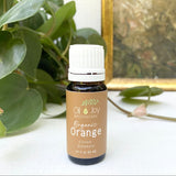 Bottle of Organic Orange essential oil from Oil Joy Apothecary on a white surface with green leaves in the background.