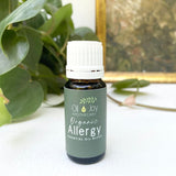 Bottle of Organic Allergy essential oil blend by Oil of Joy Apothecary on a white surface with green leaves in the background.