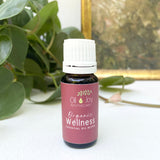 Bottle of Organic Wellness essential oil blend from Oil Joy Apothecary on a white surface with green leaves in the background.