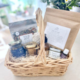 Postpartum gift basket with various items including tea, lotion, soup, bath soak, and healing products on a light surface.