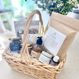 Postpartum gift basket with various items including tea, lotion, soup, bath soak, and healing products on a light surface.