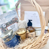 Postpartum gift basket with various items including tea, lotion, soup, bath soak, and healing products on a light surface.
