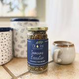 A glass jar labeled 'Immune Booster Organic Tea Blend' from Oil Joy Apothecary, placed on a kitchen counter next to a mug and with a polka dot container in the background.