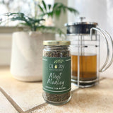 A glass jar of 'Mint Medley' organic loose leaf tea by Oil of Joy Apothecary, with a French press and a plant in the background.
