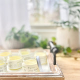 A glass jar with a metal lid labeled 'Deep Relief Rub' containing a yellowish balm, placed on a wooden surface with blurred greenery in the background.