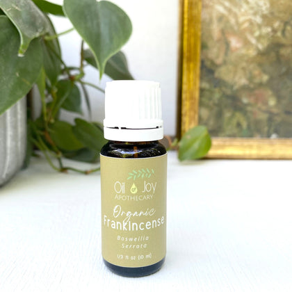 Bottle of Organic Frankincense oil by Oil Joy Apothecary on a white surface with a plant and framed picture in the background.