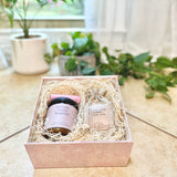 Gift set with a candle and bath soak in a decorative box on a light surface with plants in the background.